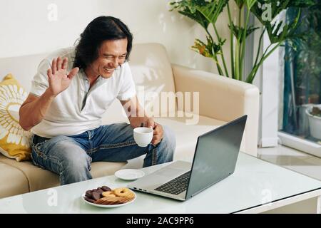 Cheerful Asian man drinking tea with cookies and video calling his friends or family members Stock Photo