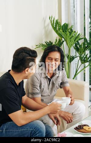 Smiling father and son drinking tea, eating cookies and discussing news at home Stock Photo