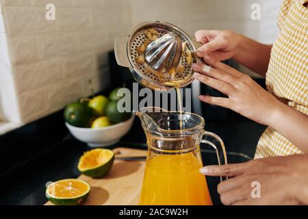 Hands of woman pouring fresh orange juice from metal manual squeezer in glass jar Stock Photo