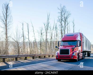 Big rig long haul red semi truck with high cab transporting commercial cargo in covered semi trailer driving on the winter wide multiline highway with Stock Photo