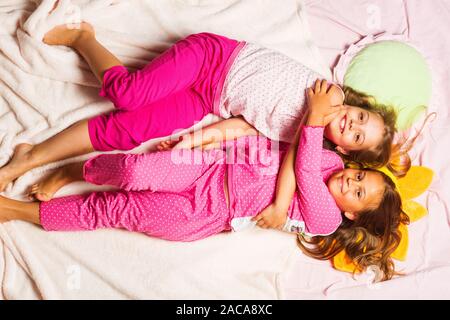 Schoolgirls have pajama party with funny pillows. Kids in pink pajamas have fun. Children with smiling faces lie on light pink blanket background and hug. Childhood, party and happiness concept. Stock Photo