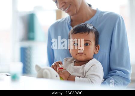 Portrait of cute baby boy sitting on mother's knees looking at camera and playing toys Stock Photo