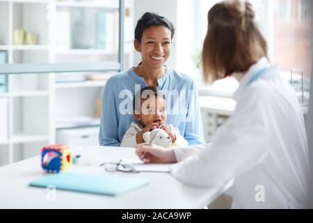 Smiling young mother sitting together with her baby and talking to the doctor during their visit at hospital Stock Photo