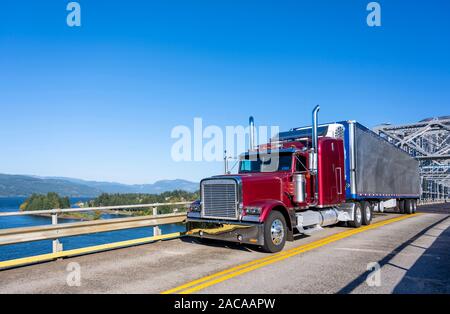 Red classic long haul big rig semi truck transporting frozen cargo in bright metal refrigerated semi trailer driving on the silver truss Bridge of God Stock Photo