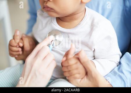 Close-up of little child sitting on mother's knees while doctor examining him with stethoscope at hospital Stock Photo