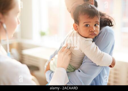 Young mother holding her little child on her hands while female doctor examining him with stethoscope at hospital Stock Photo