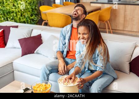 Mixed Race Marriage. Young man and woman watching comedy on tv together eating popcorn laughing cheerful Stock Photo