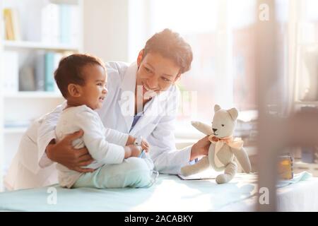 Young female doctor in white coat showing toy to her little patient and playing with him at hospital Stock Photo