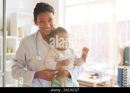 Portrait of female doctor in white coat holding baby and smiling at camera while standing at her office Stock Photo