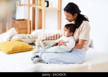 Young mother sitting on bed together with her baby and showing toy to him in the bedroom Stock Photo