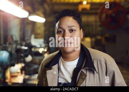 Head and shoulders portrait of contemporary mixed-race woman posing in factory workshop at looking at camera, copy space Stock Photo