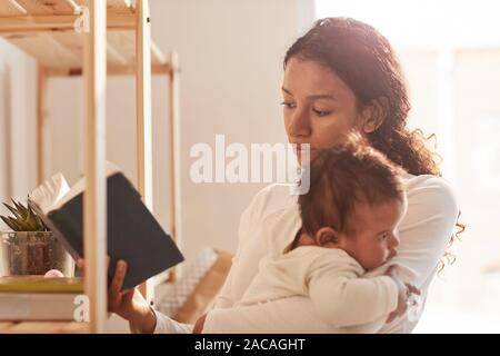 Warm-toned portrait of young African-American mother reading bonk and cuddling cute baby, copy space Stock Photo