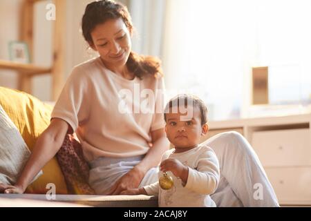 Cute child standing and holding toy while his mother sitting on sofa and playing with him at home Stock Photo
