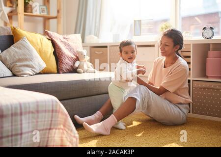 Happy young mother sitting on the floor smiling and playing with her child in the living room Stock Photo