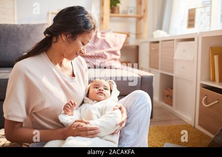 Young mother holding her baby on hands and cradling him while sitting on the floor in the room Stock Photo