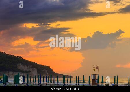 View of the bay of Peschici at sunset: sandy beach with umbrellas and trebuchet, Italy (Puglia). Peschici is famous for its seaside resorts. Stock Photo