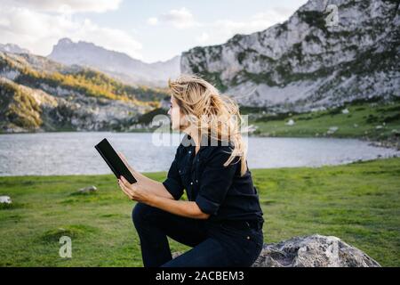 A beautiful caucasian blonde woman reads a book in a mountainous landscape with a lake Stock Photo