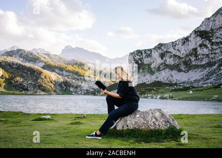 A beautiful caucasian blonde woman reads a book in a mountainous landscape with a lake Stock Photo