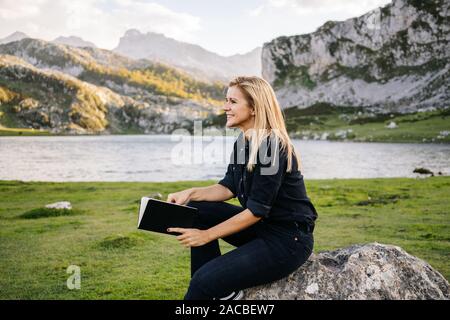 A beautiful caucasian blonde woman reads a book in a mountainous landscape with a lake Stock Photo