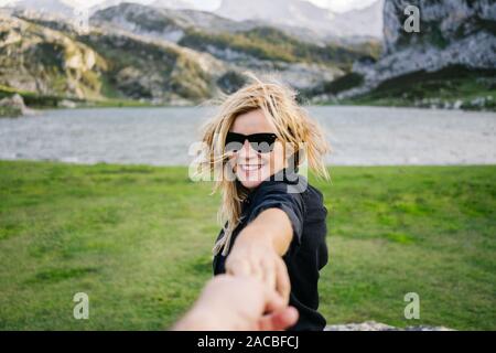 A beautiful caucasian blonde woman holds a man's hand in a mountainous landscape with lake Stock Photo