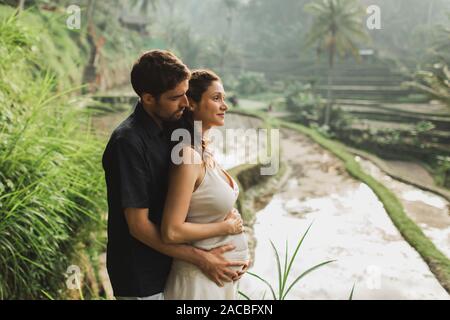 Young latin pregnant woman with husband with amazing view of Ubud rice terraces. Pregnant couple happy together. Stock Photo