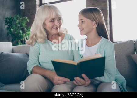 Photo of two people cute aged granny and little granddaughter sitting cozy sofa reading together learning new language teacher grandma house indoors Stock Photo