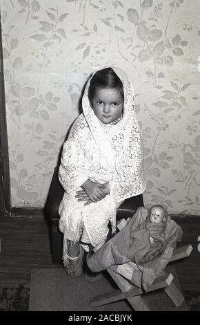 1950s, historical, nativity, inside a room a little girl in a shawl dressed as Mary, the mother of Jesus, sitting on a chair and beside her, a little doll as baby jesus lying in a small wooden cradle, England, UK. Stock Photo