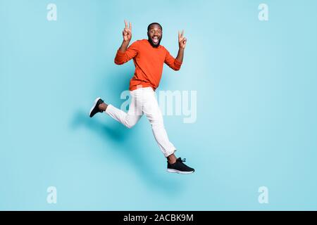Full length body size photo of cheerful positive casual man running jumping mixed-race wearing white pants trousers sneakers showing double v-sign Stock Photo