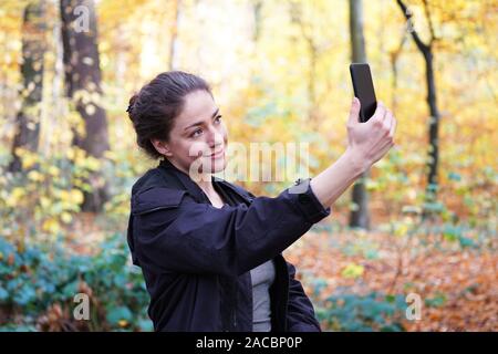 young woman in her 20s taking selfie self portrait with her smartphone mobile phone during autumn walk in forest Stock Photo