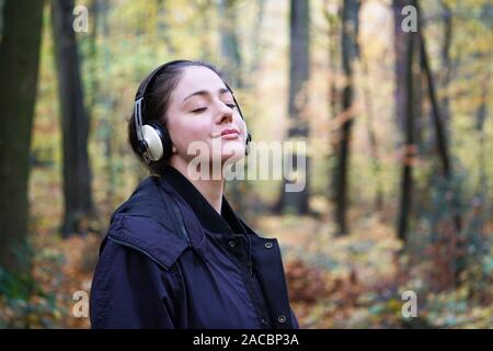young woman in her 20s listening to music with wireless headphones in forest - candid outdoor lifestyle in autumn - with copy space