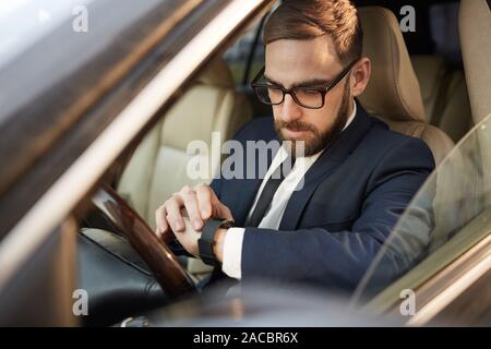 Serious young businessman in eyeglasses sitting in the car and looking at his watch on his hand Stock Photo