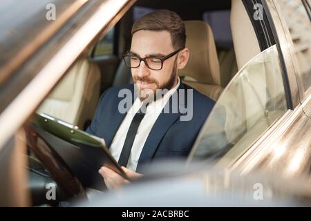 Young businessman in eyeglasses reading documents and smiling while sitting in his car Stock Photo