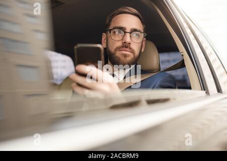 Serious young businessman using his mobile phone and looking at view of the city while sitting in the car Stock Photo