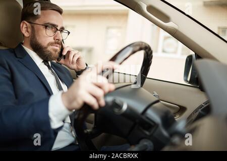 Young serious businessman in eyeglasses talking on mobile phone and driving the car Stock Photo