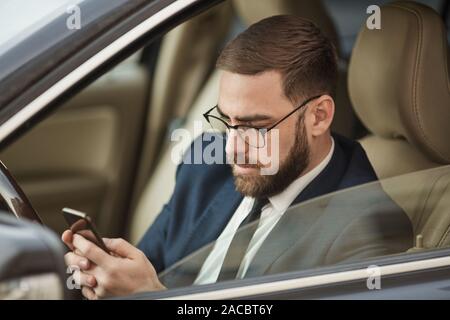 Young man in eyeglasses sitting in the car and texting message on mobile phone Stock Photo
