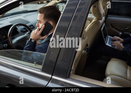 Young businessman talking on the phone and driving the car with other businessman sitting on back seat and working on laptop Stock Photo