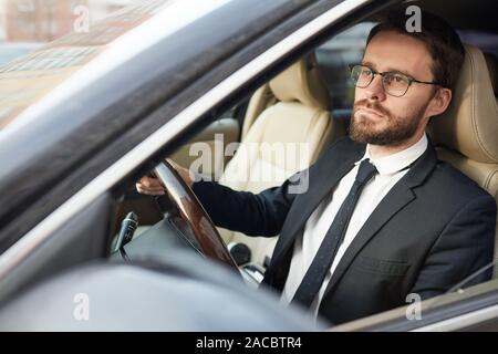 Serious young bearded businessman in eyeglasses sitting behind the wheel and driving the car he concentrating on the road Stock Photo