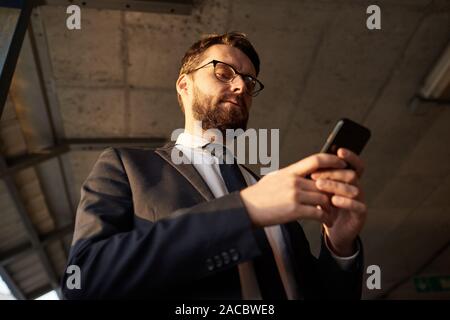 Low angle view of bearded businessman in suit standing and working online using his mobile phone Stock Photo