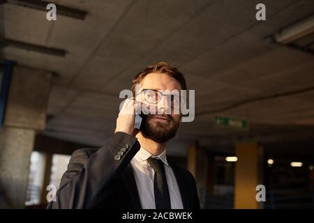 Young bearded businessman wearing eyeglasses and suit using mobile phone in the parking Stock Photo