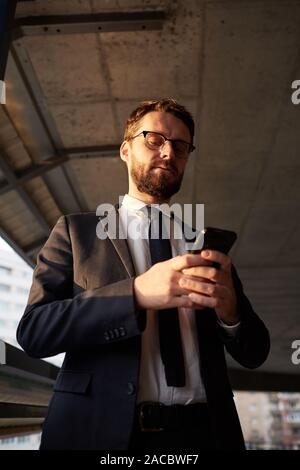 Young serious businessman wearing eyeglasses and black suit standing outdoors and typing e message on his mobile phone Stock Photo