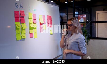 Project management agile methodology, concept. A young blonde woman in glasses, gluing colored sticky notes on a magnetic Board. Stock Photo