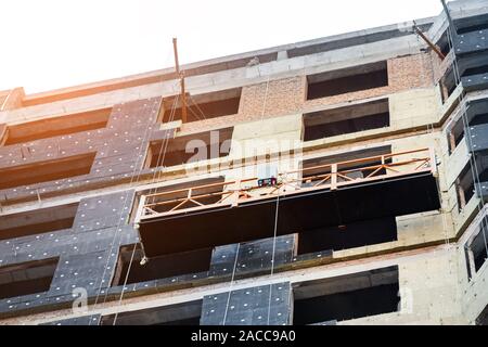 Suspended construction craddle near wall of hightower residentaial building with insulation and ventilated facade on construction site. Engineering Stock Photo