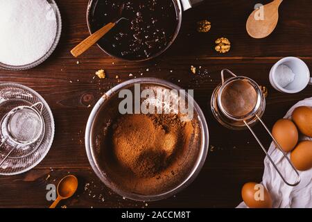 Sifted cocoa powder and flour in metal bowl next to melted dark chocolate, walnuts and eggs ingredients for delicious homemade brownie cake on wooden Stock Photo