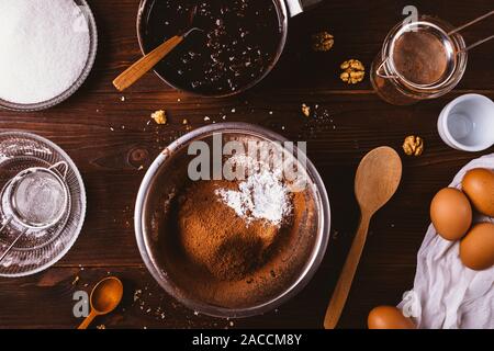 Metal bowl with sifted cocoa powder and flour next to melted dark chocolate, walnuts and eggs ingredients for delicious homemade brownie cake on woode Stock Photo