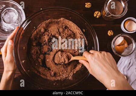 Woman's hands mixing melted chocolate and cocoa powder in large bowl to make dough for delicious brownie cake on dark wooden table next to ingredients Stock Photo