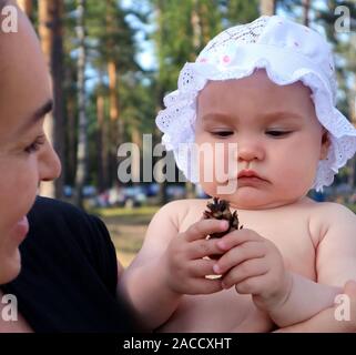 Adorable baby girl dressed in a cap with ruffles playing with pine cone outdoors Stock Photo