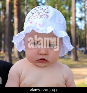 Adorable pensive baby girl dressed in a cap with ruffles outdoors  against pine forest Stock Photo