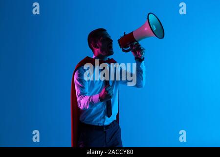 Businessman dressed like superhero shouting by megaphone on blue background Stock Photo