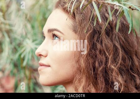 Portrait of girl with brown curly hair on background of green bush sea buckthorn, close-up face Stock Photo