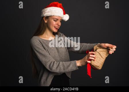 A beautiful girl in a Santa Claus hat on a dark gray background opens a Christmas gift and breaks the Kraft gift packaging. The blonde model tears up Stock Photo
